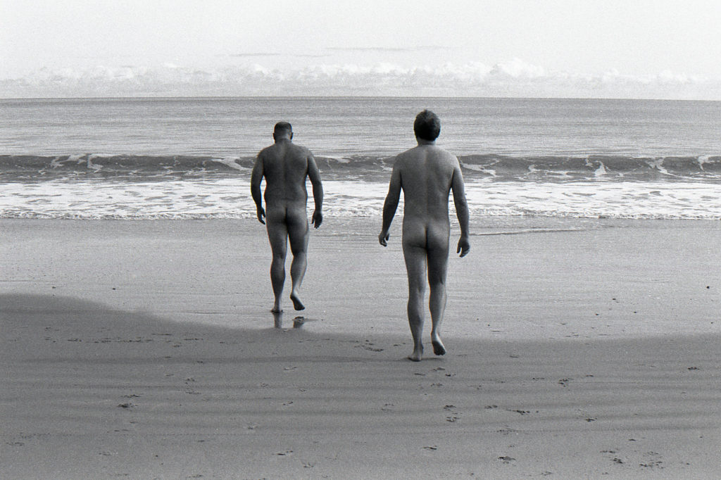 Black and white image of two nude men walking on the beach away from the camera towards the ocean.